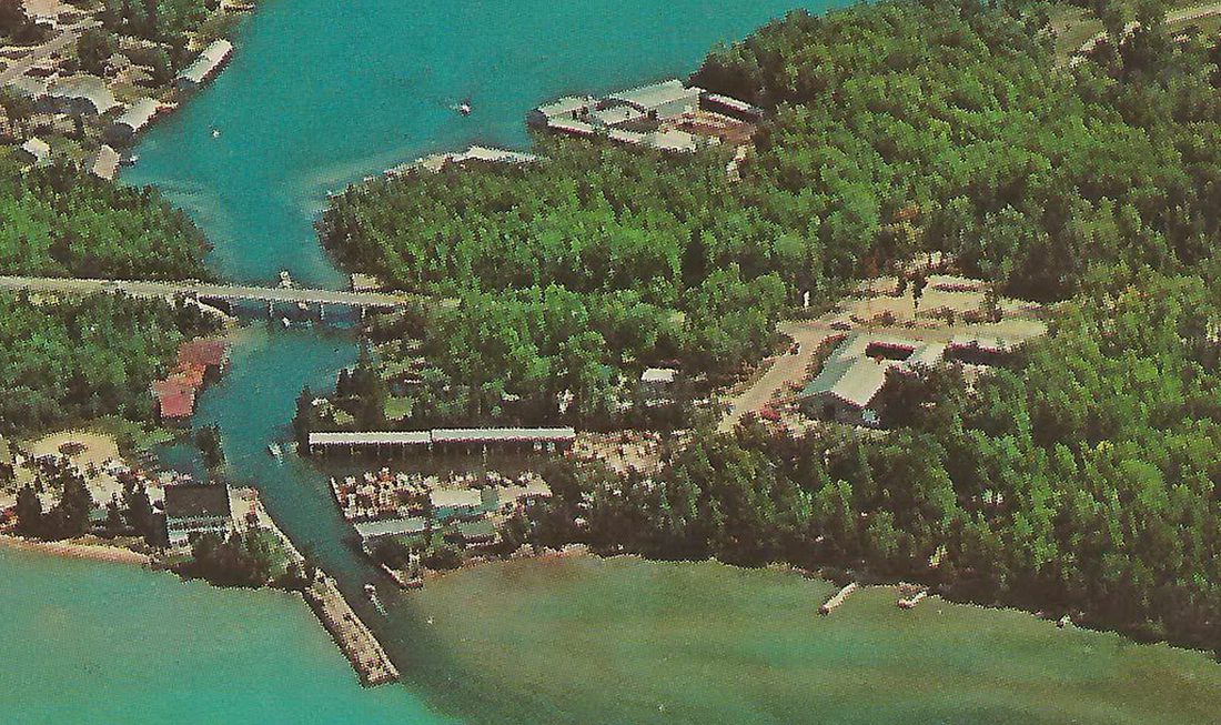 1950s aerial postcard: the Clam River crossing under a multi-pier bridge, boathouses and a long white marina building on the banks, boats in the channel, a breakwater reaching into turquoise Torch Lake.