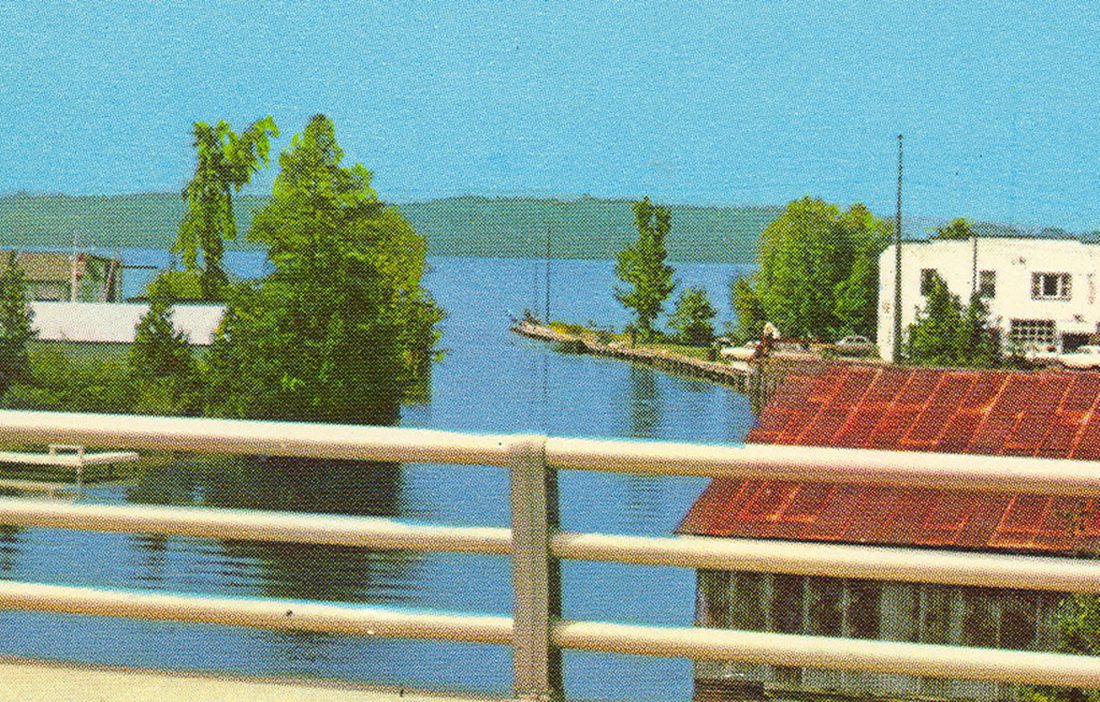 Mid-century tinted postcard from the Clam River bridge deck: a two-story white store and boathouse at the river mouth, piling docks into the channel, Torch Lake beyond.